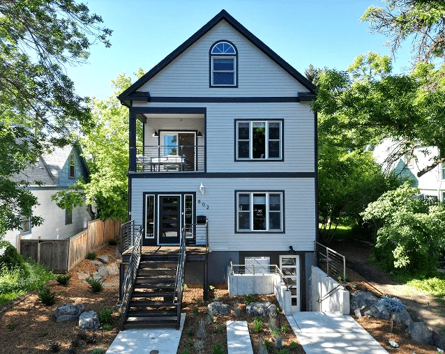 A modern, three-story house with black trim sits amid lush greenery, featuring a welcoming front porch and landscaped garden area.