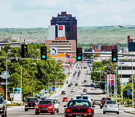 Busy urban street with multiple cars, green traffic lights, American flag, and tall buildings under a partly cloudy sky.