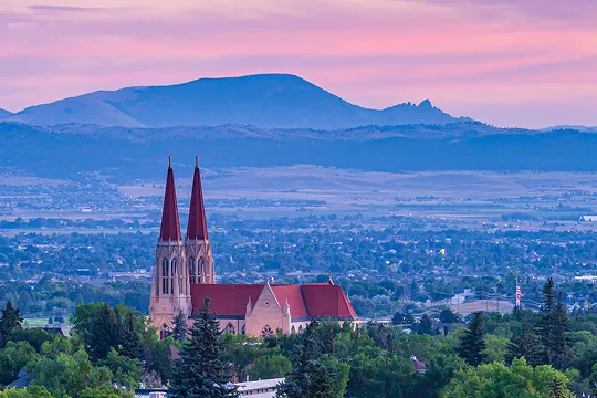 View of a church with twin red spires amidst trees, cityscape, and mountains under a pink and purple sunset sky.