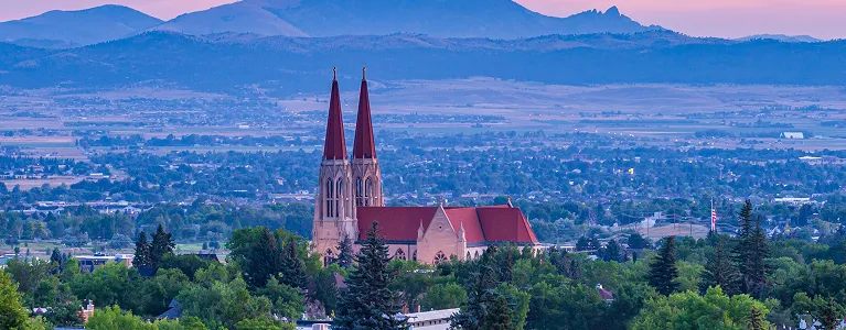 Panoramic view of a large church with two red steeples surrounded by greenery and distant mountains at sunset.