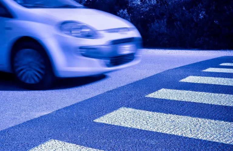 Blurred white car approaching a pedestrian crosswalk on an asphalt road at night.
