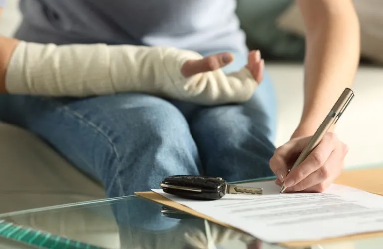 Person with a bandaged arm signing a document on a glass table with car keys nearby.