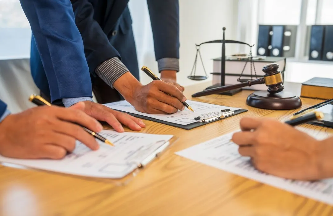 Three individuals in formal attire reviewing and signing legal documents at a wooden desk.