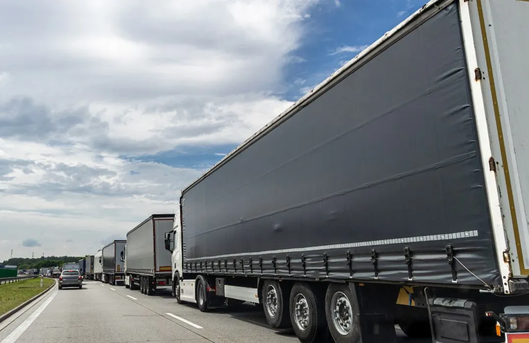 Line of large cargo trucks with dark trailers on highway under partly cloudy sky.