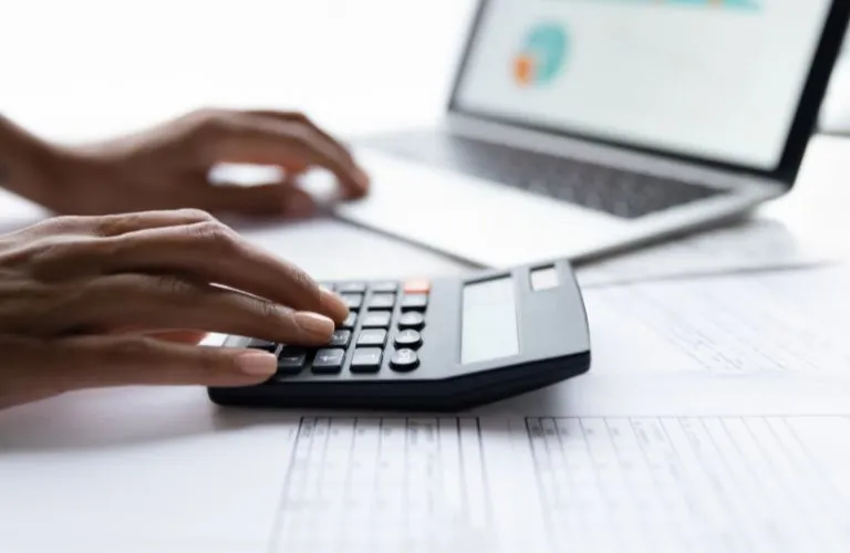 Person using a black calculator and laptop on a desk with financial documents.