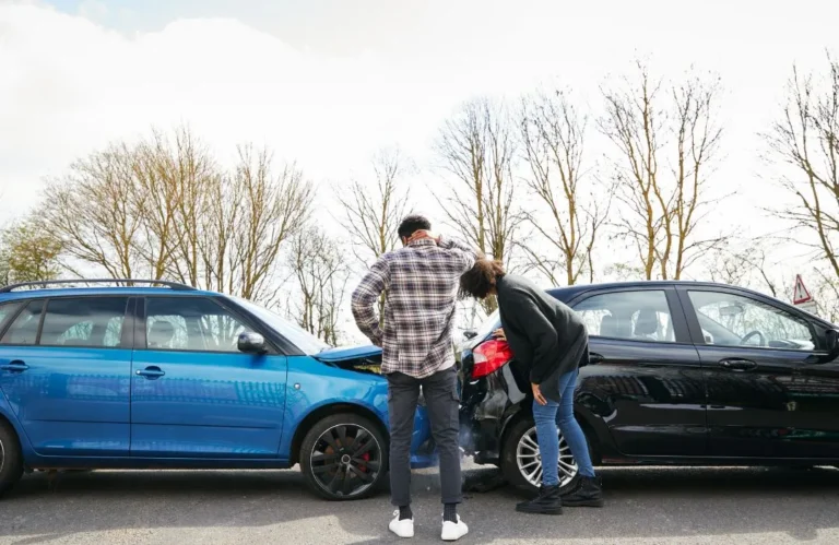 Two individuals inspecting damage after a rear-end collision between a blue and black car on a road.