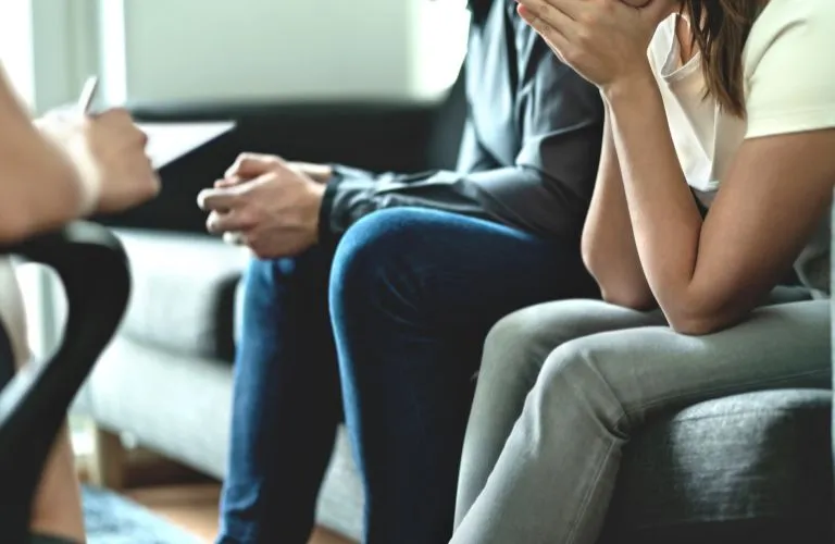 Two individuals seated on a gray couch during a counseling session, one covering their face.