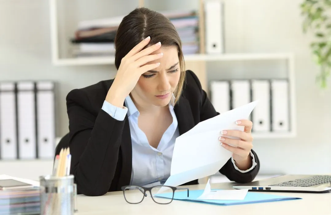 Professional woman in black blazer reviewing document at desk with laptop and glasses.