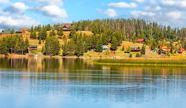Lakeside view of forested hillside with scattered cabins under a partly cloudy sky.