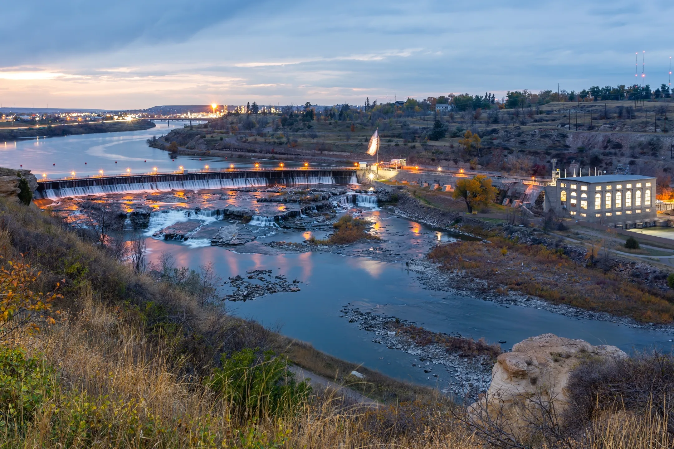 Dusk view of a river dam with cascading water, illuminated buildings, and surrounding autumn vegetation.
