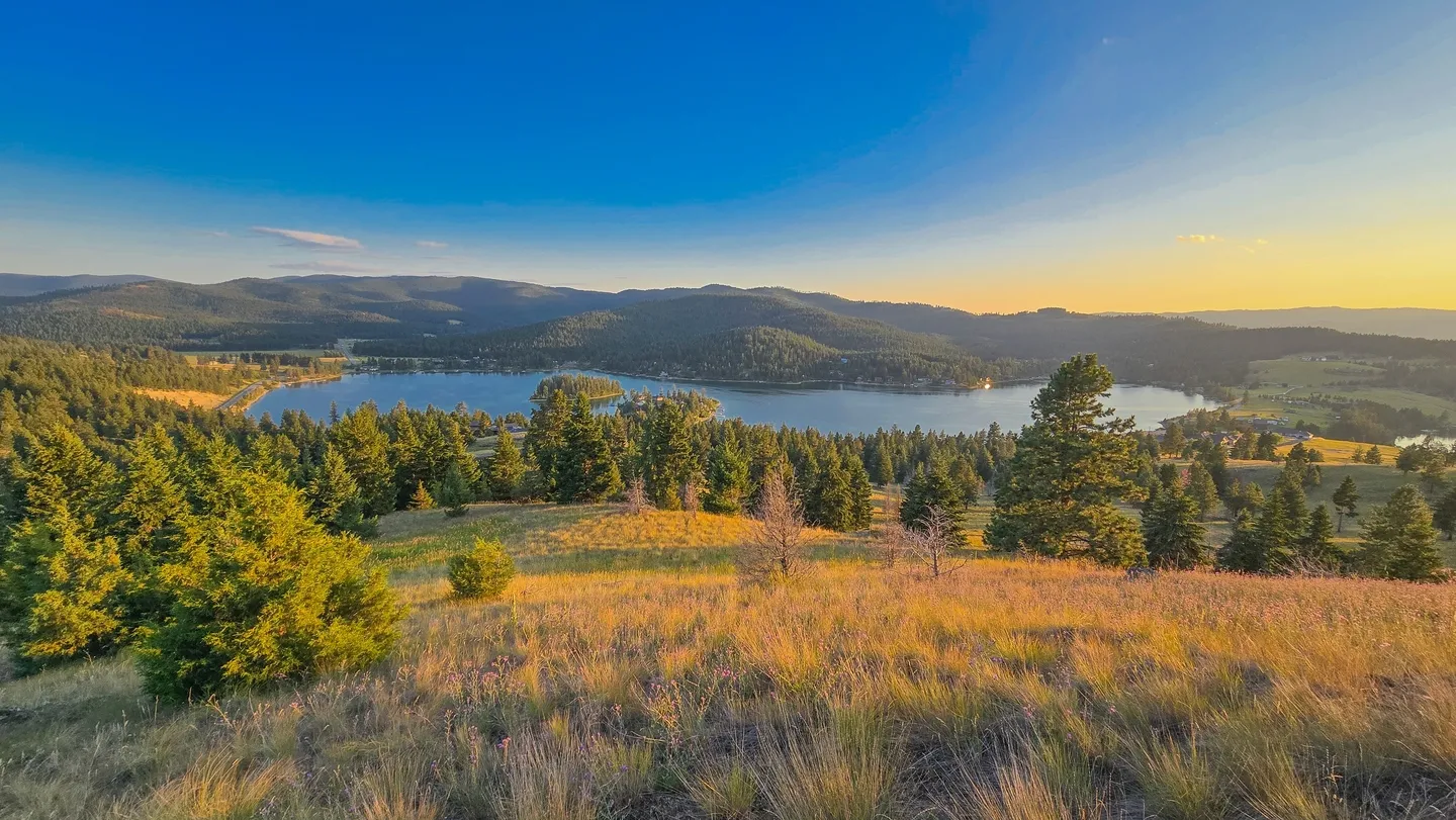 Panoramic view of a lake surrounded by forested hills and grassy fields under a clear blue sky at sunset.