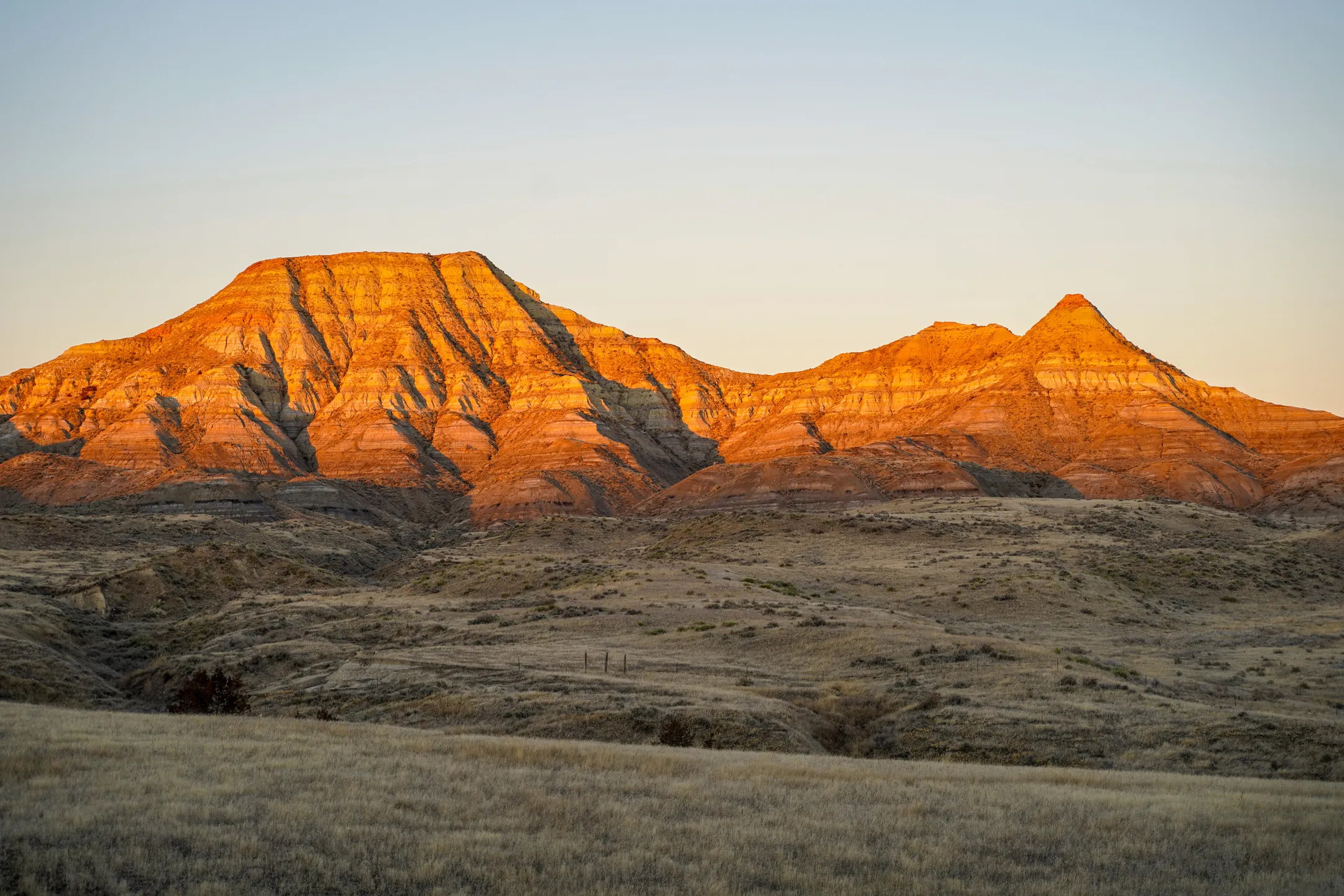 Sunlit layered rock formations and dry grassland under a clear sky at sunset.