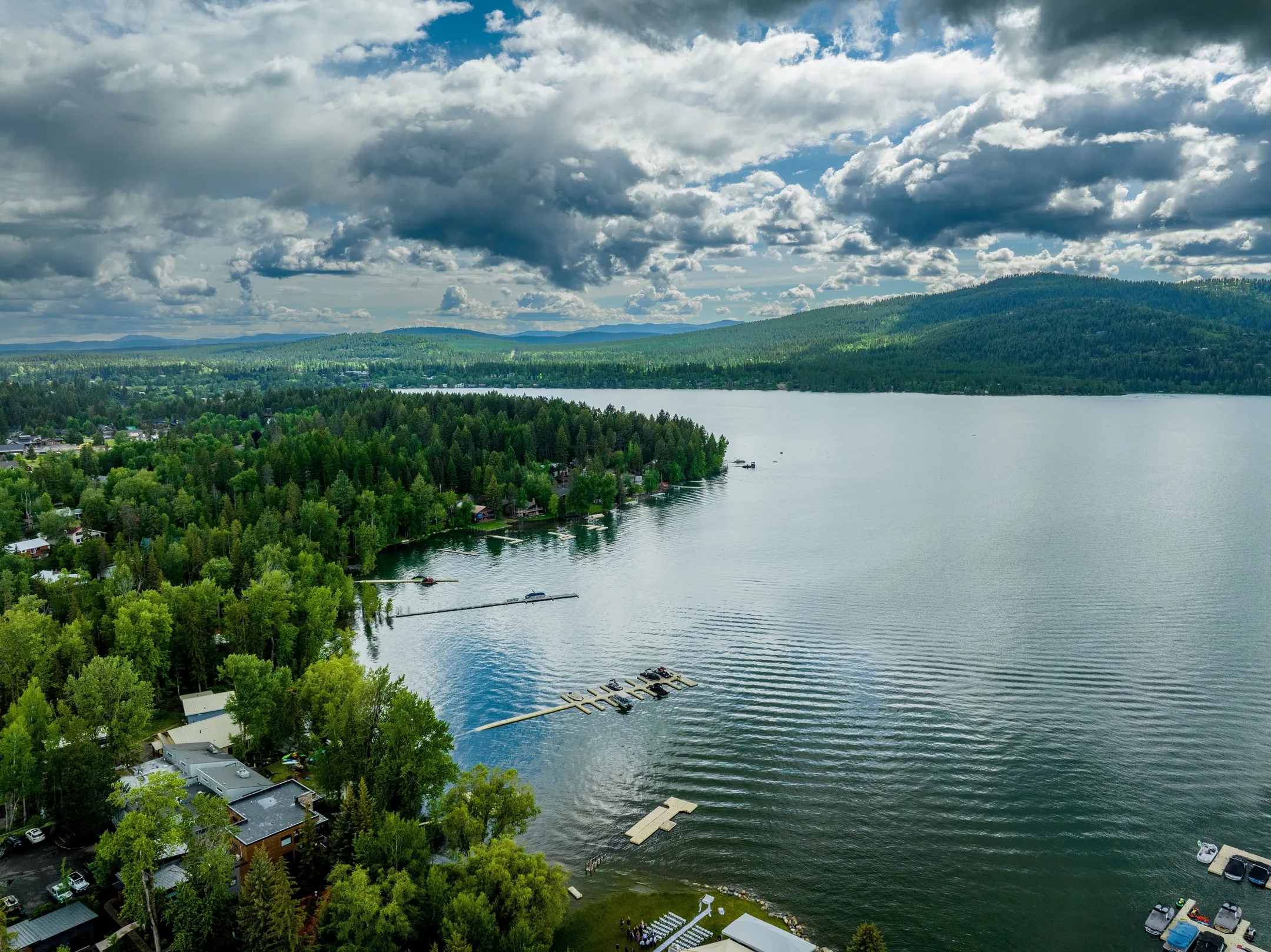 Aerial view of a large lake surrounded by dense green forest, docks, and scattered buildings under a cloudy sky.